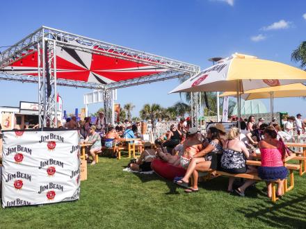 Festival goers sitting under shaded tents at a sponsor booth at Sand in My Boots Festival